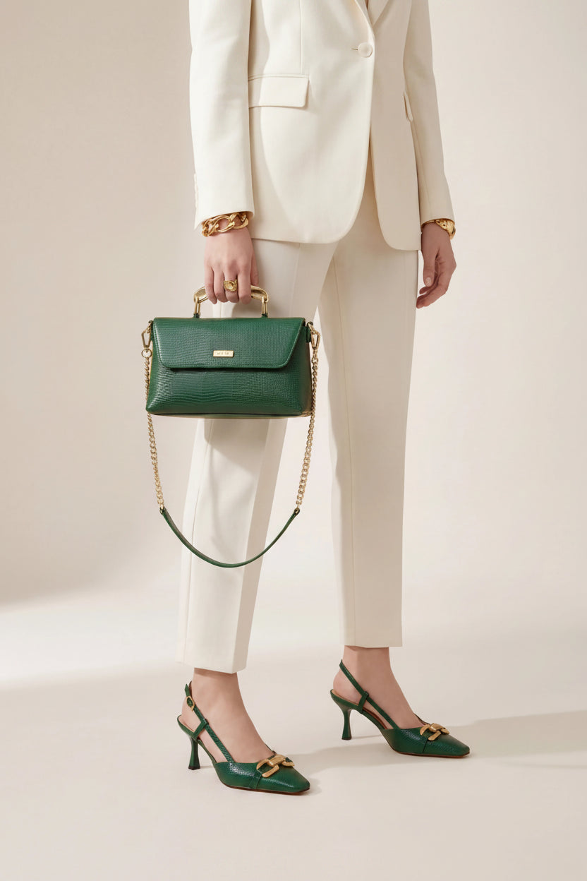 A model wearing a cream-colored blazer and trousers holds a dark green structured handbag with a gold chain strap and green pointed-toe heels with gold hardware.