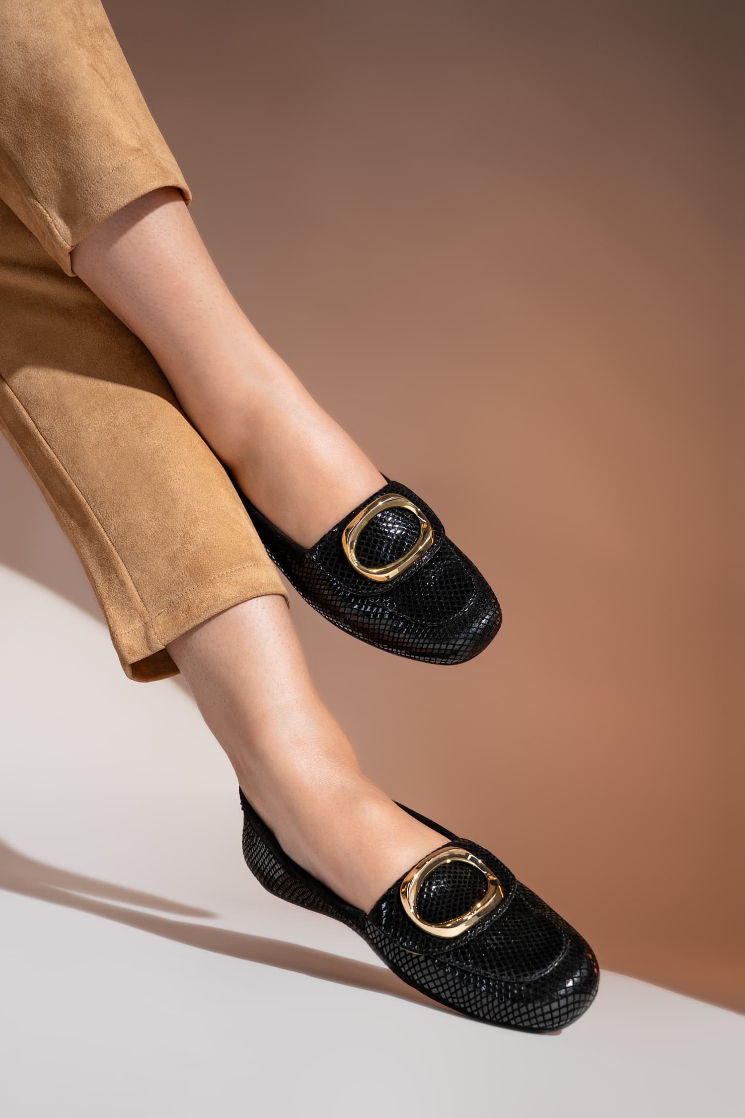 Black loafers with gold buckles worn by a person on a beige background