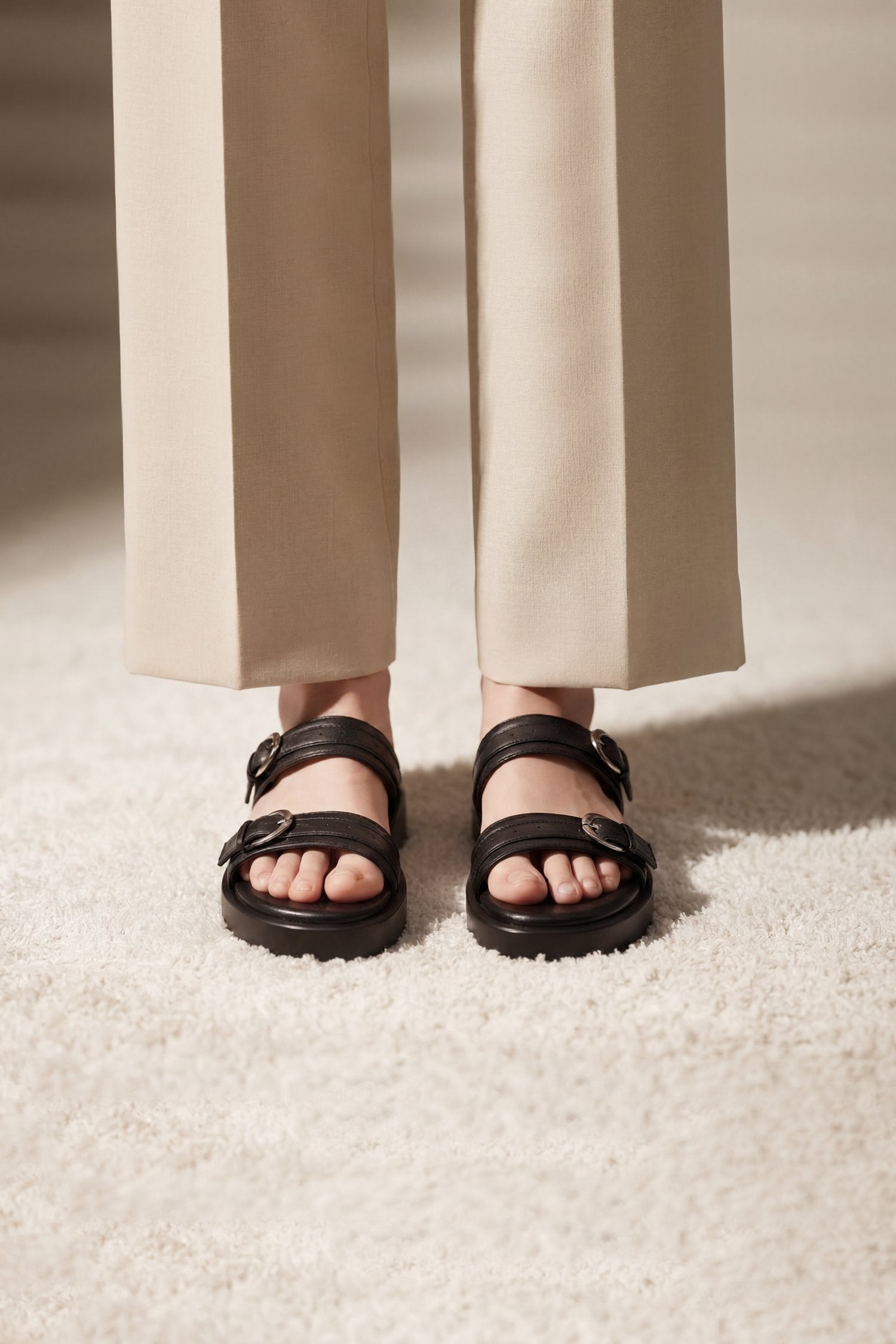 Black double-strap leather sandals with buckle details, shown on a model's feet wearing beige trousers on a textured cream rug.