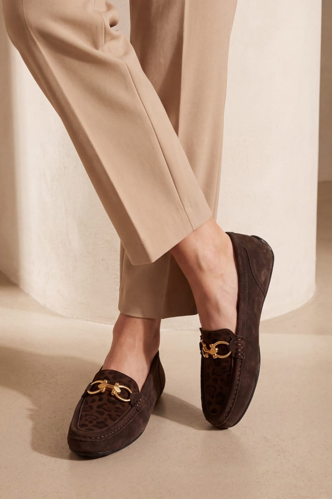 A person wearing dark brown suede and leopard-print loafers with gold horsebit hardware, paired with beige trousers, standing against a neutral beige background.