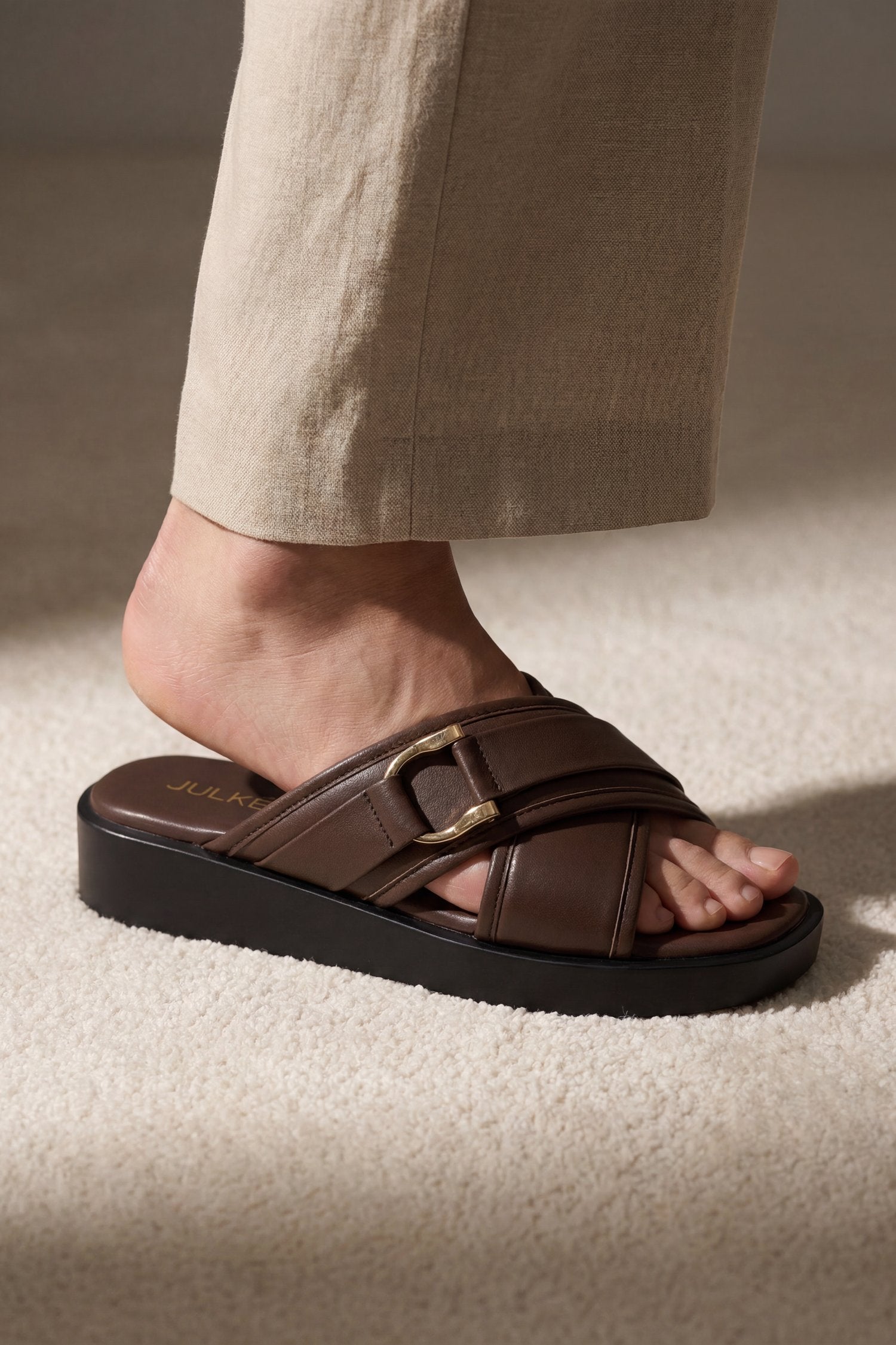 A close-up of a person's foot wearing a dark brown leather slide sandal with a wide strap, a gold-tone buckle, and a thick black platform sole, paired with beige trousers on a textured beige carpet.