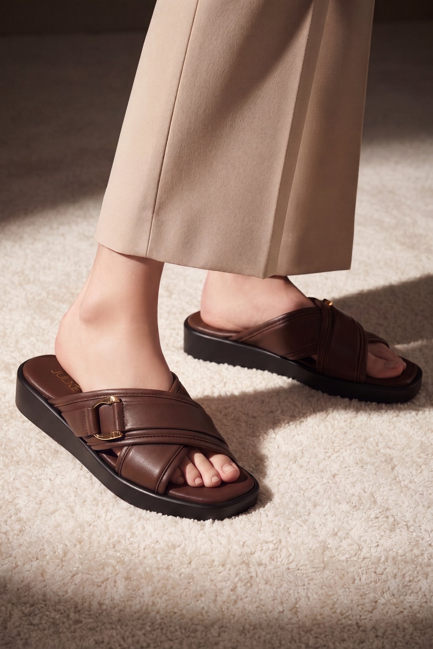 A person wearing dark brown leather slide sandals with wide straps and a buckle detail, paired with beige trousers, standing on a textured beige carpet.