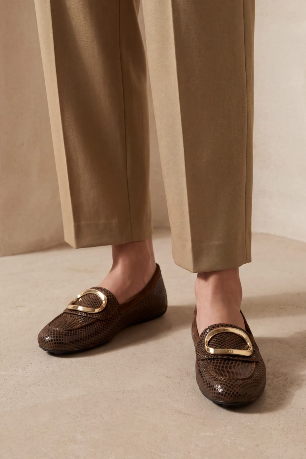 A person wearing brown snakeskin-patterned loafers with large gold-tone O-shaped metal accents, paired with straight-leg beige trousers, standing on a neutral beige floor.