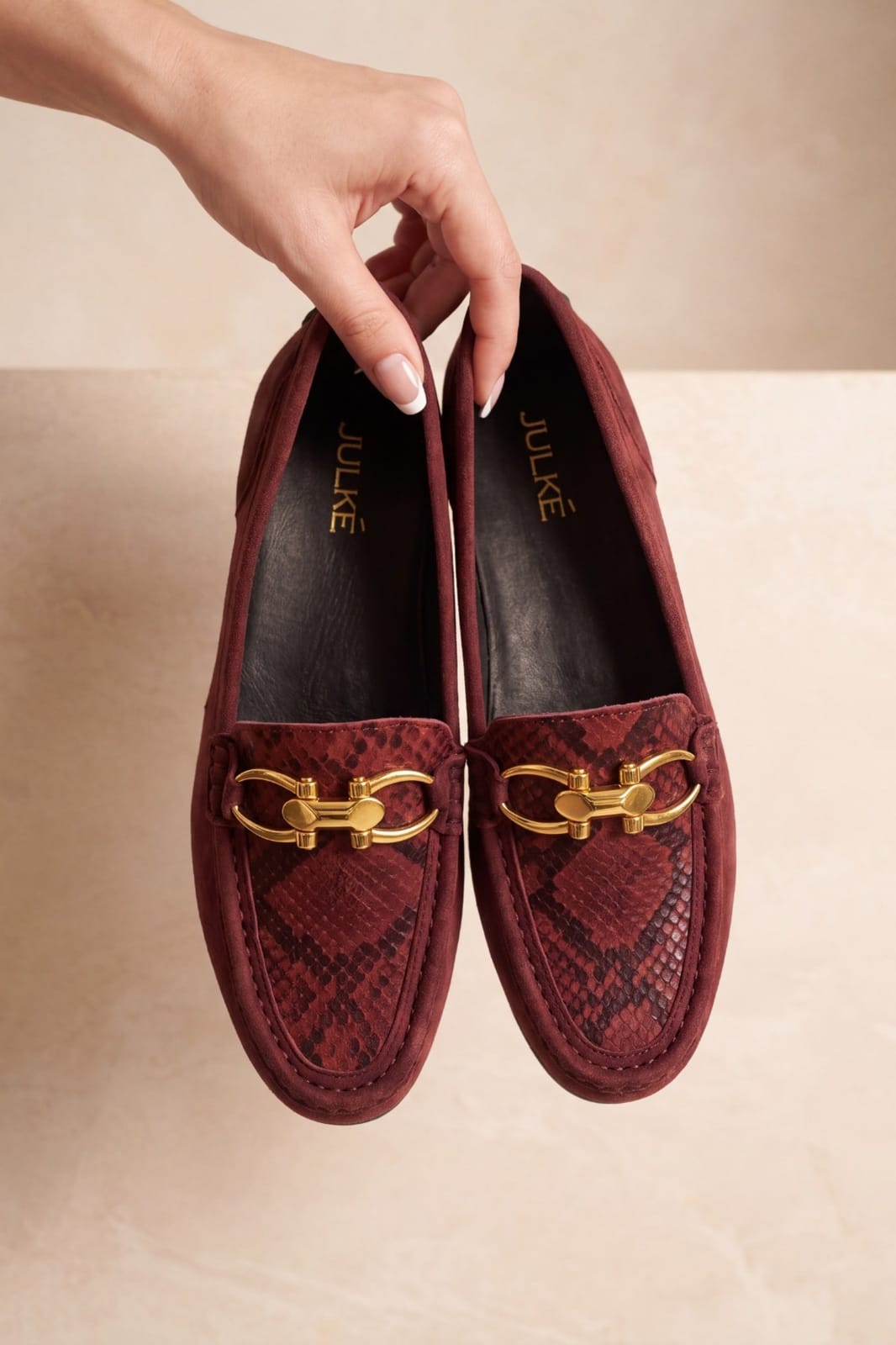 A pair of burgundy snakeskin-patterned loafers with gold-tone horsebit hardware, held by a hand against a neutral beige background. The brand name 'JULIE' is visible on the insoles.
