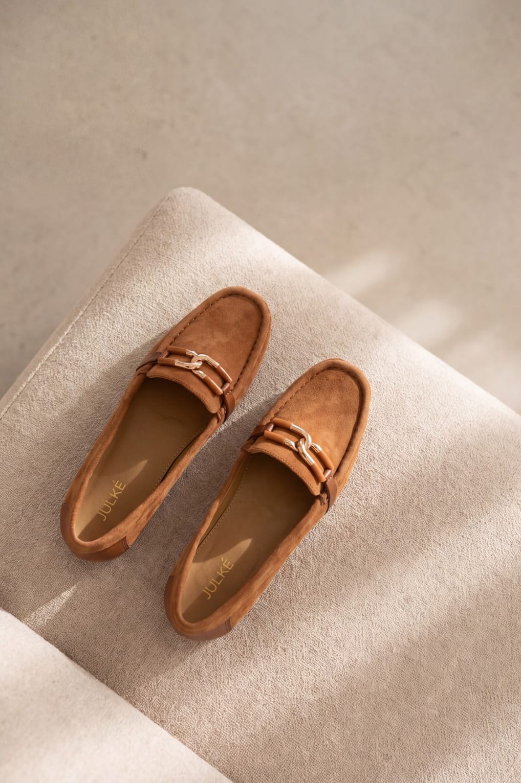 A pair of tan suede loafers with gold-tone horsebit straps, displayed on a textured beige fabric cushion.