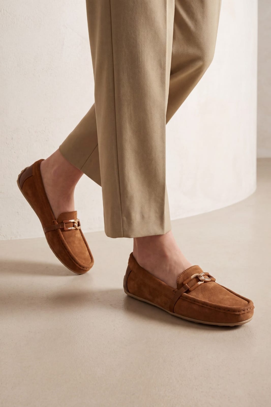 A person wearing tan trousers and brown suede loafers with a small gold-tone buckle detail, shown in a clean indoor setting with a light beige floor and wall.