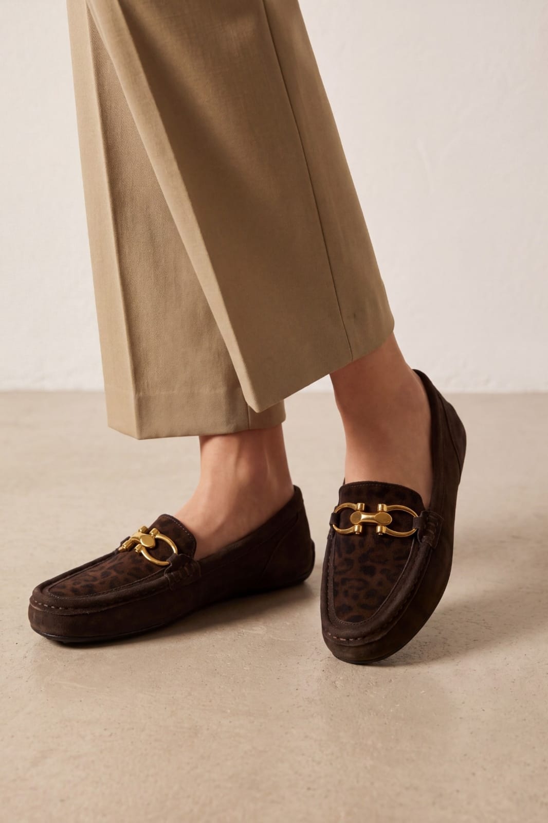 A person wearing brown loafers with a leopard print pattern and a gold horsebit detail, paired with beige wide-leg trousers, standing on a light beige floor against a neutral wall.