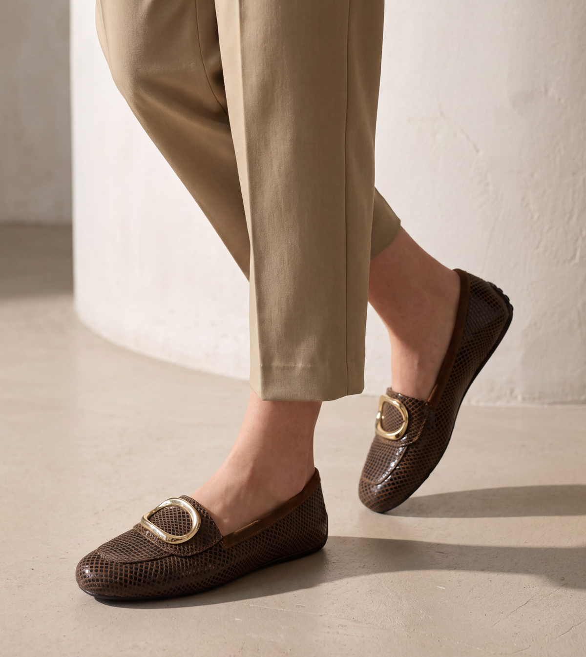 A person wearing dark brown textured loafers with gold-tone oval hardware and beige cropped trousers, standing on a light concrete floor in a minimalist indoor setting.