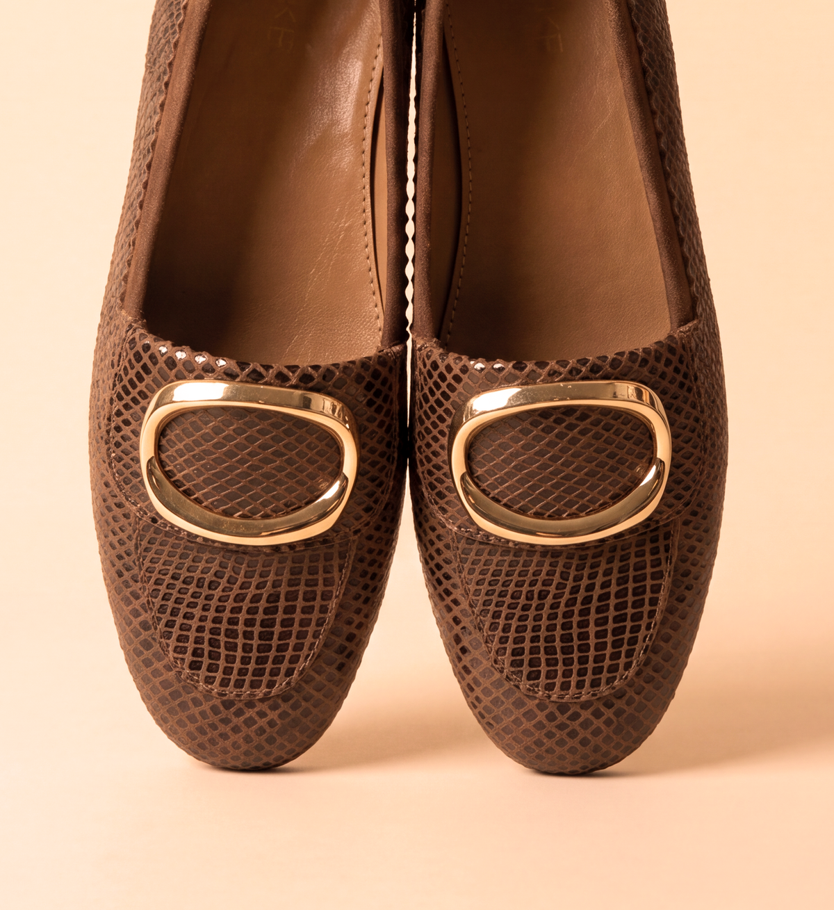 A pair of brown snakeskin-patterned loafers with large gold-tone oval buckles, shown from the front on a solid beige background.